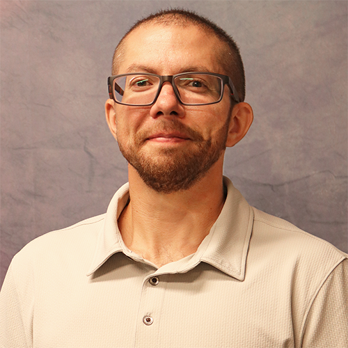 Portrait of a man with short hair and a trimmed beard wearing rectangular glasses and a light polo shirt, facing the camera against a gray backdrop.