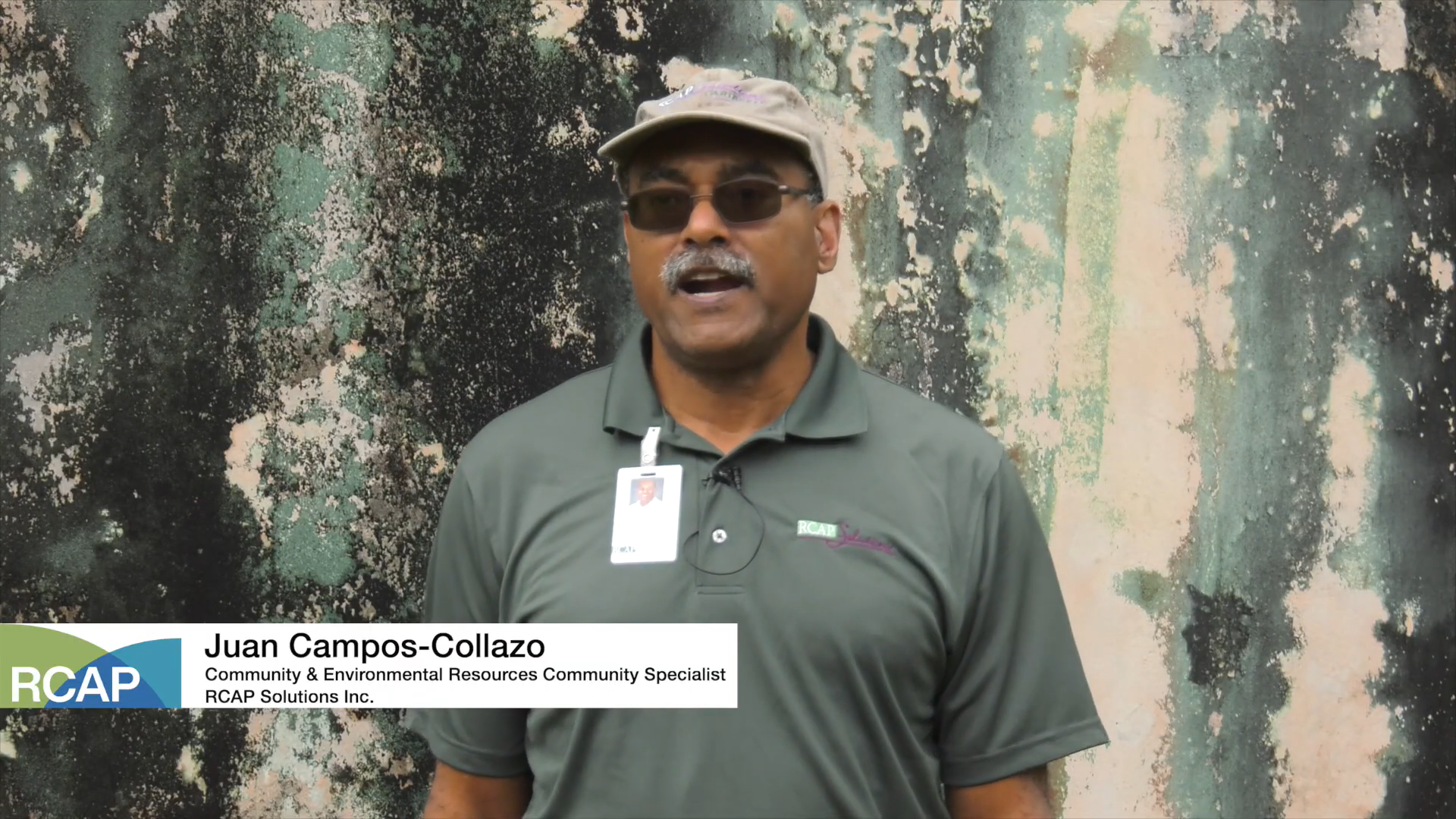 Man in a green polo with an ID badge speaks to the camera against a mottled wall.
